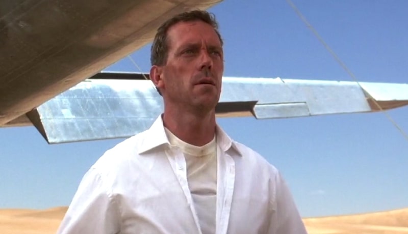 Hugh Laurie in a button-up white shirt in the desert near a broken down airplane.