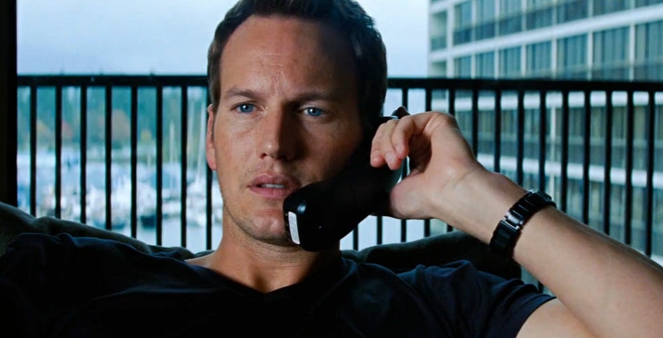 Patrick Wilson holding a phone to his head with a stern look near a balcony.