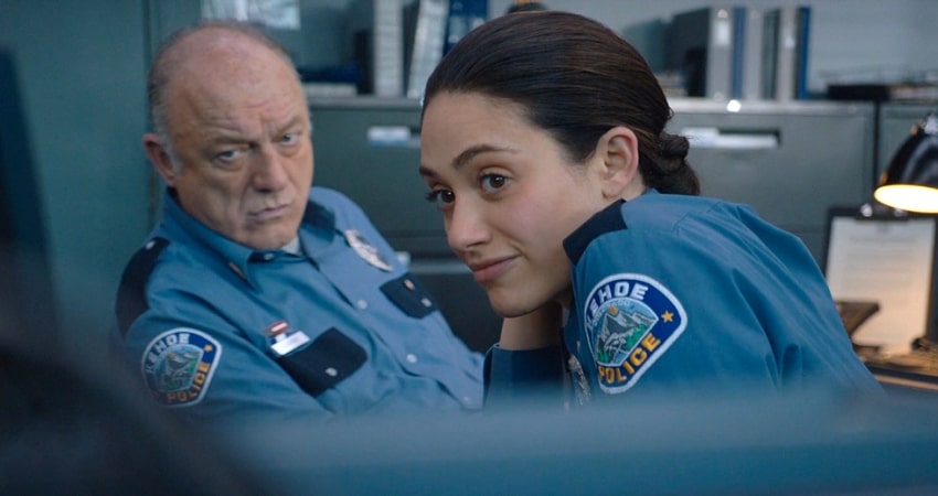 Emmy Rossum with an old cop smiling with Kehoe cop uniforms in an office in Cold Pursuit.