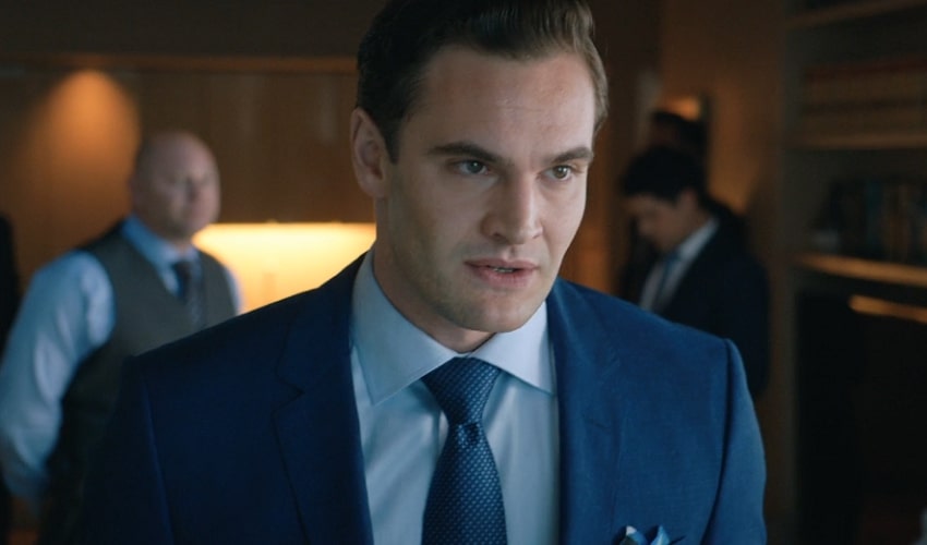 Tom Bateman in a blue suit and tie with people behind him.