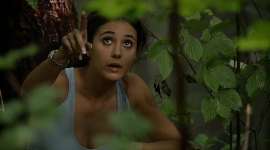 Emmanuelle Chriqui hiding in a forest behind leaves and pointing to the sky in Wrong Turn.