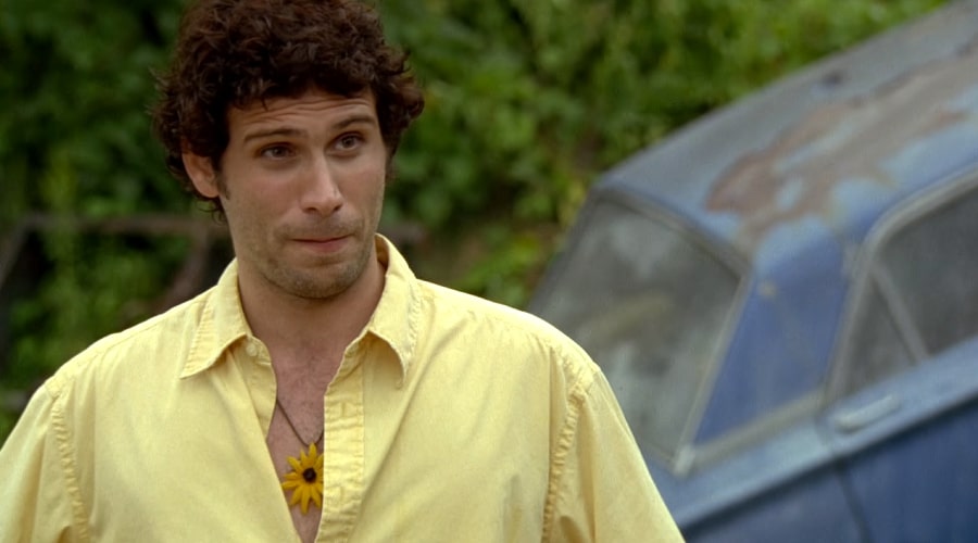 Jeremy Sisto with a flower necklace and yellow short near an abandoned vehicle.