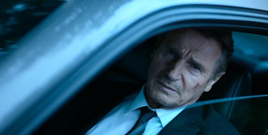 Liam Neeson in a suit and tie with a scar on his cheek inside a silver car in Blacklight.